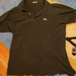 Svart pikétröja från Lacoste - Svart pikétröja från Lacoste med klassisk krage och två knappar framtill. Tröjan har korta ärmar och den ikoniska gröna krokodilloggan broderad på bröstet. Materialet är mjuk bomull som känns skönt mot huden. Perfekt för dig som gillar stilren och enkel design.På bilderna ser den lite vit ut men de är den inte i verkligen geten utan den är i helt nyskick och helt svart som den ska.skulle säga att den passar dig om du är 175-180 kanske 