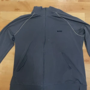 Mörkblå ziptröja från BOSS - Snygg mörkblå ziptröja från BOSS med diskret logga på bröstet. Tröjan har lång ärm, dragkedja hela vägen och praktiska fickor framtill. Perfekt för dig som gillar stilrena och sportiga plagg.