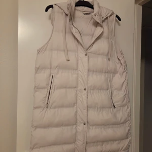 Ljusbeige lång dunväst med huva - Säljer en ljusbeige, quiltad dunväst med huva och snörning. Västen är lång, har två fickor med dragkedja och stängs med både dragkedja och tryckknappar. Perfekt för lager-på-lager och chill stil under kyliga dagar.