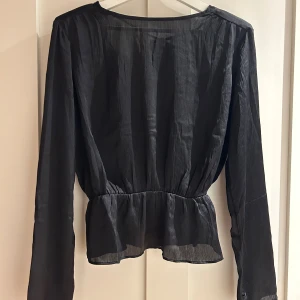 Svart omlottblus med knyt från Bik Bok - Säljer en svart omlottblus från Bik Bok i tunn, lätt skimrande polyester. Blusen har v-ringning, knytband i nacken, markerad midja och volang nedtill. Långa ärmar med små knappar vid ärmslut. Perfekt för dig som gillar stilrena och trendiga plagg.