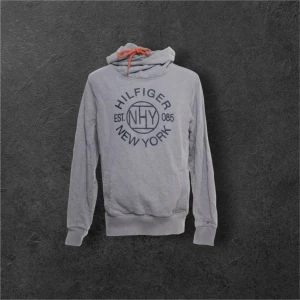 Grå hoodie Tommy Hilfiger L - Snygg grå hoodie från Tommy Hilfiger i storlek L med vintage fit. Tröjan har tryck med Hilfiger New York på bröstet, orange snörning i huvan och ribbade muddar. Tillverkad i mjuk bomull med lite elastan för extra komfort. Perfekt för chill dagar.