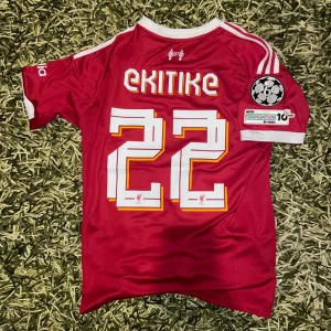  Ekitike #22 Liverpool 25/26 fotbollströja - Röd Liverpool fotbollströja med Ekitike och nummer 22 på ryggen. Kortärmad modell med vita detaljer och tryck, Champions League-märke på ärmen och Standard Chartered-logga på bröstet. Slim fit i lätt, ventilerande polyester. Perfekt för dig som älskar LFC.