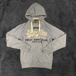 Blå hoodie från Superdry - Snygg grå hoodie från Superdry i storlek XS med vintage-tryck på bröstet och kontrasterande vita snören. Tröjan har huva, känguruficka och ribbade muddar vid ärmar och nederkant. Perfekt för chill dagar och streetstyle.