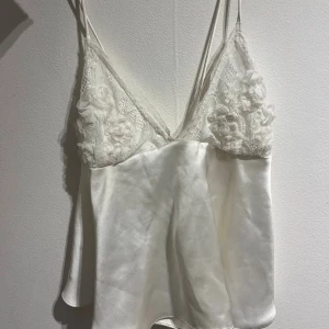 Vit siden linnetopp med spetsdetaljer - Supersöt vit linnetopp i glansigt siden med tunna axelband och spets upptill. Spetsen har broderade blommor som ger en romantisk vibe. Toppen är v-ringad och har en flowy passform som känns både lyxig och chill. Perfekt för dig som gillar mjuka material och fina detaljer.