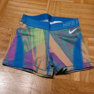 Nike Pro färgglada träningstights-shorts XS - Nike Pro shorts i XS med ett galet färgglatt, randigt mönster i blått, grönt, rosa och beige. De har en bred blå midja med Nike-logga och är gjorda i stretchigt syntetmaterial som sitter tight och bekvämt. Perfekta för gymmet eller sport.
