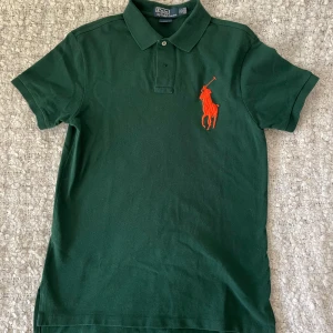 Grön pikétröja från Polo Ralph Lauren - Snygg grön pikétröja från Polo Ralph Lauren med orange broderad polospelare på bröstet. Tröjan har klassisk krage, korta ärmar och knappar vid halsen. Custom fit-modell i mjuk bomull som är skön att bära. Perfekt för dig som gillar stilrena och sportiga plagg.