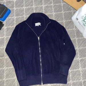 Marinblå stickad kofta från Maison Margiela - Säljer en marinblå stickad kofta från Maison Margiela med ribbad struktur och hel dragkedja framtill. Koftan har hög krage och långa ärmar, perfekt för dig som gillar stilrena och exklusiva plagg. Tillverkad i Italien och har Margielas ikoniska vita sömmar bak i nacken. Perfekt för höst å vinter, den håller en varm 🤟🏽🤟🏽🤟🏽