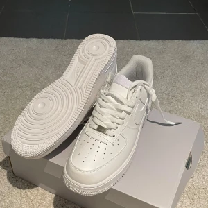 Nike Air Force 1 helvita sneakers - Helt nya Nike Air Force 1 ’07 i klassiskt helvitt utförande, storlek 44. Skorna är helt oanvända och kommer i originalkartong. Den ikoniska modellen med tidlös design, slitstark ovandel i läder och den välkända Air-sulan som ger komfort hela dagen. Perfekta både till vardags och för en stilren streetlook.
