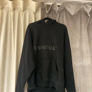 Svart Essentials hoodie med huva - Svart hoodie från Essentials med stor ficka framtill och tryckt logga över bröstet. Tröjan har huva och är tillverkad i mjuk bomull och polyesterblandning, perfekt för chill dagar. Oversized passform och långa ärmar ger en avslappnad vibe.