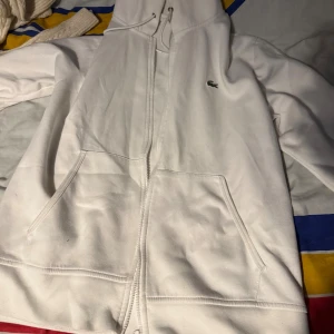 Vit hoodie från Lacoste med dragkedja - Vit hoodie från Lacoste med klassisk krokodillogga på bröstet. Tröjan har huva med snörning, två fickor framtill och hel dragkedja. Perfekt för chill dagar och enkel att matcha med jeans eller joggers.