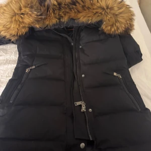 Svart pufferjacka från Hollies - Säljer en svart pufferjacka från Hollies med stor fluffig huva och snygg pälsdetalj i brunt. Jackan har två snedställda fickor med dragkedja och kraftig dragkedja framtill. Perfekt för kalla dagar och har Arctic Expedition-tryck på insidan.