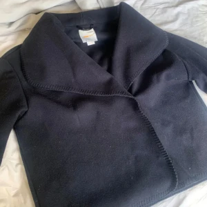 Svart kappa från Gina Tricot - Snygg svart kappa från Gina Tricot med bred krage och markerade sömmar längs kanterna. Jackan har en rak och stilren form, långärmad och tillverkad i mjukt material som känns skönt mot huden. Perfekt för dig som gillar klassisk och enkel stil.