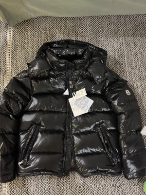 Svart Moncler pufferjacka size 1 - Säljer en svart pufferjacka från Moncler med glansig finish och klassisk logga på ärmen. Jackan har huva, två snedställda fickor med dragkedja och kraftig YKK Vislon-dragkedja framtill. Perfekt för kalla dagar och riktigt snygg till streetwear-stilen. Fits S, unisex.
