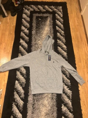 Grå hoodie från Polo Ralph Lauren - Snygg grå hoodie från Polo Ralph Lauren med klassisk logga på bröstet. Tröjan har huva med snörning, ribbade muddar och en stor magficka. Perfekt för chill dagar och enkel att matcha med jeans eller joggers.