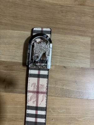 Burberry rutig bälte med silverdetalj - Snyggt bälte från Burberry med klassiskt rutigt mönster i beige, svart och rött. Bältet har en unik silverspänne med Burberrys ikoniska riddare. Tillverkad i skinn och levereras med originalpåse och box. Perfekt accessoar för att lyfta din outfit.