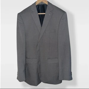 Grå kavaj med diskret mönster - Snygg grå kavaj med diskret småmönstrad yta och klassisk passform. Kavajen har två framfickor med lock, bröstficka och fyra knappar vid ärmslutet. Fodret är mörkblått och känns lyxigt mot huden. Perfekt för dig som vill ha en stilren look.