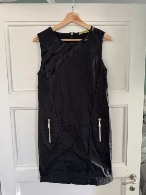 Svart ärmlös klänning Versace Jeans - Svart kortklänning från Versace Jeans med snygga guldfärgade dragkedjor och coola meshdetaljer upptill. Klänningen är ärmlös och har en rak, figurnära passform. Materialet är bomull och dragkedjorna har märkets ikoniska logga.