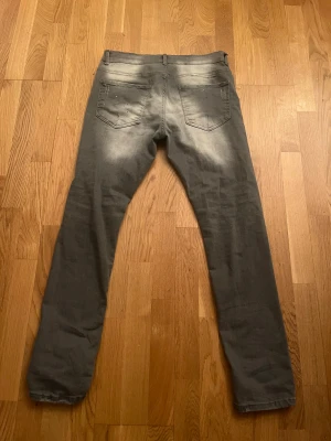 Grå Dsquared2 jeans med färgstänk - Snygga grå jeans från Dsquared2 med coola vita och blå färgstänk och slitningar på framsidan. Modellen har fem fickor, knappgylf och klassisk röd Dsquared2-tag vid gylfen. Jeansen har en rak passform och är tillverkade i jeansmaterial.