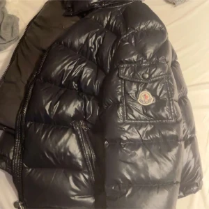 Svart pufferjacka från Moncler - Säljer en svart pufferjacka från Moncler med glansig finish och klassisk logga på ärmen. Jackan har dragkedja framtill, fickor med lock och quiltad design. Perfekt för kalla dagar och har en riktigt snygg streetstil.