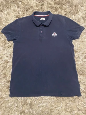 Mörkblå pikétröja från Moncler - Snygg mörkblå pikétröja från Moncler med klassisk krage och två knappar framtill. Tröjan har Moncler-logga broderad på bröstet och är tillverkad i mjuk bomull. Kortärmad och med normal passform, perfekt för en clean och stilren look.