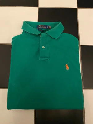 Polo Ralph Lauren piké - Snygg grön pikétröja från Polo Ralph Lauren med klassisk krage och två knappar framtill. Tröjan har den ikoniska orangea loggan broderad på bröstet. Perfekt för en stilren och sportig look.