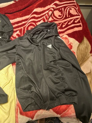 Svart Adidas vindjacka med huva - Svart vindjacka från Adidas med klassiska vita ränder längs ärmarna och logga på bröstet. Jackan har huva, dragkedja framtill och två sidofickor. Materialet är lätt och syntetiskt, perfekt för att hålla vinden ute. Snygg och sportig look med normal passform.