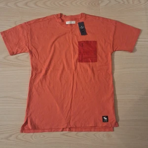 Orange t-shirt Abercrombie med ficka stl 15-16 år - Snygg orange t-shirt från Abercrombie med korta ärmar och en cool bröstficka med dragkedja. Tillverkad i mjuk bomull och har en liten logotyp längst ner. Perfekt för dig som gillar streetwear och vill sticka ut med färg.