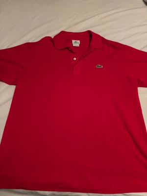 Röd pikétröja från Lacoste - Snygg röd pikétröja från Lacoste med klassisk krage och två knappar framtill. Tröjan har det ikoniska gröna krokodilmärket broderat på bröstet. Perfekt för dig som gillar stilren och sportig look.