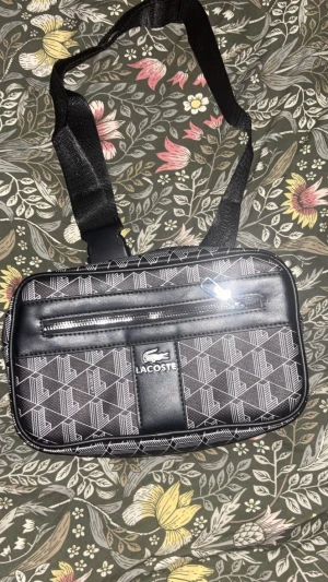 Svart väska från Lacoste - Snygg svart axelväska från Lacoste med geometriskt mönster i vitt. Väskan har justerbar rem, dragkedja framtill och ett stort huvudfack. Materialet är syntet med detaljer i skinn-look. Perfekt för att bära dina essentials och ge din outfit en cool touch.