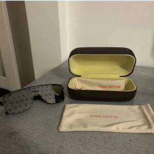 Louis Vuitton monogram solglasögon - Snygga svarta solglasögon från Louis Vuitton med spegelglas och ikoniskt monogrammönster över hela linsen. Sköldformad modell med bred båge och diskreta silvriga detaljer. Kommer med originalfodral och putsduk.
