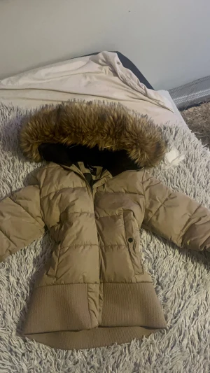 Beige pufferjacka med fuskpäls - Säljer en beige pufferjacka med stor huva kantad av fuskpäls. Jackan har ribbade muddar vid ärmslut och nederkant, dragkedja framtill och en ficka med knapp. Perfekt för kalla dagar och jätte mysig att bära💞 den är xs men passar s lika bra! skicka gärna prisförslag btw jag postar alltid samma dag eller andra!