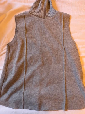 Grå stickad tröja med polokrage - Snygg grå stickad väst med hög polokrage och ribbad struktur. Västen är ärmlös och har en avslappnad passform, perfekt att slänga över en skjorta eller t-shirt för en cool lager-på-lager-look.