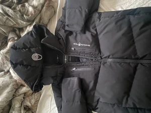 Svart dunjacka Sail Racing 170 - Säljer en svart dunjacka från Sail Racing med huva och feta Sail Antarctica Expedition-detaljer. Jackan har två dragkedjeförsedda bröstfickor och är fylld med Allied Feather & Down 500 fill power. Perfekt för kalla vinterdagar och riktigt snygg streetstil.