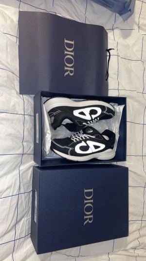Dior B30 svarta och vita sneakers skor - Säljer ett par Dior B30 sneakers i svart och vitt med vit sula. Skorna har mesh och syntetmaterial, snörning och en modern, sportig look med tydlig Dior-logga på sidan och plösen. Perfekta för dig som gillar exklusiva sneakers med statement.       Dom passar bra till alla outfits och pris går att diskuteras
