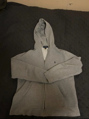 Grå hoodie från Polo Ralph Lauren XL - Snygg grå hoodie från Polo Ralph Lauren i storlek XS. Tröjan har huva, dragkedja framtill och två fickor. Klassisk Polo-logga broderad på bröstet. Mjuk insida i fleece och långärmad modell, perfekt för chill dagar.