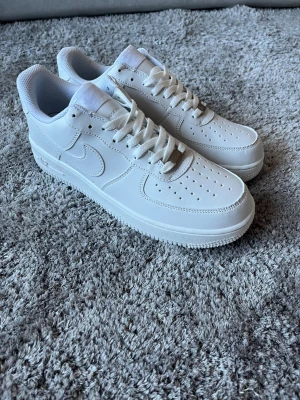 Nike Air Force 1 helvita sneakers - Nike Air Force 1 sneakers i helvit färg med klassisk låg siluett. Skorna har snörning, perforerad tå och rundad sula. Tillverkade i skinn med vit mesh på insidan för extra komfort. Snyggt broderad Nike Air-logga på hälen och metallplatta på snörningen. Inte äkta