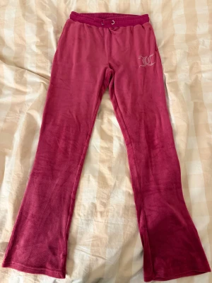 Juicy Couture rosa velourbyxor - Juicy Couture velourbyxor i en snygg mörkrosa färg med bootcut-ben och snörning i midjan. Byxorna har bakfickor och ett glittrigt JC-logo på höften i strass. Perfekta för en chill och trendig look.