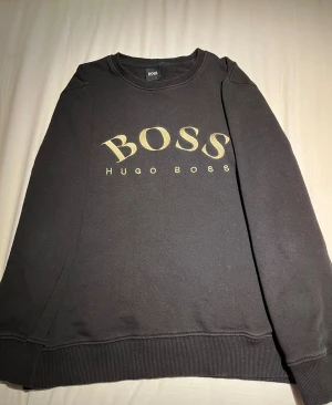 Svart sweatshirt från Hugo Boss - Svart sweatshirt från Hugo Boss med stort guldigt BOSS-tryck på bröstet. Klassisk rund halsringning, ribbade muddar och lång ärm.  Storlek S. Frågor om plagget eller om du vill ha mer bilder så är det bara att skriva! Priset kan diskuteras