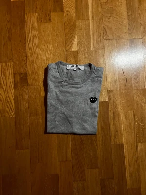 Grå Comme des Garçons Play t-shirt - Säljer en grå t-shirt från Comme des Garçons Play med det ikoniska svarta hjärta med ögon broderat på bröstet. Klassisk rund hals och mjukt bomullstyg som känns skönt mot huden. Sitter rätt så liten i storleken. Hör av dig vid funderingar!  