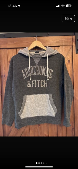 Cool unik Grå hoodie Abercrombie & Fitch  - Mysig mörkgrå hoodie från Abercrombie & Fitch i storlek XS. Tröjan har ljusgrå huva och magficka, snörning vid halsen och stort tryck framtill. Relaxed fit och tillverkad i mjuk bomullsmix, perfekt för chill dagar. Relaxed fit sp kan passa vissa som bär S. Typ av slitning vid slutet av atmarna och längst ner på hoodien ( de ska vara så ) som får den att se använd ut.