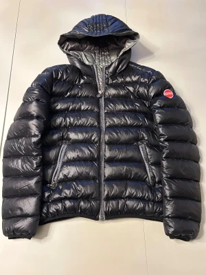 Svart Colmar dunjacka med huva - Snygg svart dunjacka från Colmar med glansig finish, vadderad design och huva. Jackan har två dragkedjeförsedda fickor, tydlig logga på ärmen och grov dragkedja framtill. Perfekt för kalla dagar när du vill hålla stilen. Skick som ny, använda cirka 3 gånger