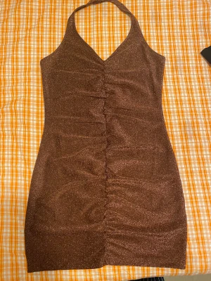 Glittrig brun halterneck klänning - Supersnygg brun glittrig halterneck klänning. Kort modell med åtsittande passform och rynkade detaljer framtill. Perfekt för dig som vill sticka ut med en glittrig look. Materialet är stretchigt och skimrar i ljuset.