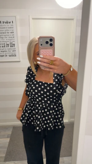 Svart prickig blus med volanger - Säljer en svart blus med vita prickar och volangdetaljer på axlarna. Toppen har en fyrkantig ringning, knytband i ryggen och är ärmlös. Materialet känns lätt och flowy, perfekt för dig som gillar statement-plagg med retrovibe.