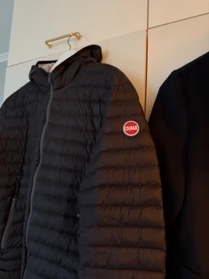 Svart pufferjacka från Colmar (52) - Säljer en svart pufferjacka från Colmar med huva och dragkedja. Jackan har ett quiltat mönster och Colmar-logga på ärmen. Tillverkad i syntetmaterial och perfekt för kyliga dagar. Snygg och enkel att matcha till olika outfits.