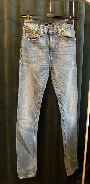 Blå jeans från Nudie Jeans Co - Snygga ljusblå jeans från Nudie Jeans Co med klassisk femficksdesign och orangea sömmar. Jeansen har rak passform, normal midja och är tillverkade i mjukt bomullsmaterial. Baksidan har Nudie Jeans broderi på fickorna och en brun läderpatch vid linningen. Str W27 L32 pris 299kr, kan diskuteras.