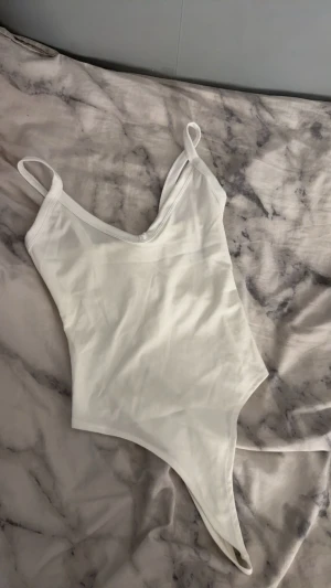 Vit body med smala band - Säljer en stilren vit body med smala axelband och v-ringad hals. Bodyn är ärmlös och har en enkel, clean design som är lätt att matcha med olika outfits. Materialet känns mjukt och lätt, perfekt för varma dagar.