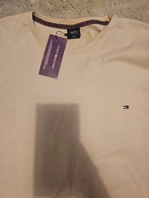 Beige t-shirt  - Snygg beige t-shirt i  storlek 3XL. Klassisk rund hals och korta ärmar. Diskret Hilfiger-logga broderad på bröstet och coolt mönstrad kant på insidan av halsen. Mjuk bomull som känns skön mot huden. Perfekt för dig som gillar stilrena plagg. Vet inte om den är äkta