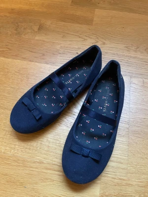 Mörkblå ballerinaskor med rosett - Snygga mörkblå ballerinaskor från Holly & Whyte med rund tå och platt sula. Skorna har en söt rosett framtill och ett band över foten. Insidan är dekorerad med ett gulligt körsbärsmönster i rosa och grönt. Yttermaterialet är i textil och sulan i syntet.