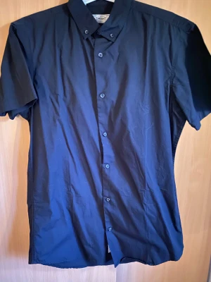 Mörkblå kortärmad skjorta - Snygg mörkblå skjorta från The Product med klassisk krage och korta ärmar. Skjortan har knappar hela vägen framtill och är tillverkad i mjuk bomull som känns skön mot huden. Perfekt för dig som gillar enkel och stilren design.