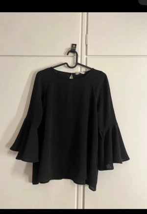 Svart blus med volangärmar - Säljer en svart blus med vida volangärmar och rund halsringning. Blusen har en lätt och flowy passform, perfekt för dig som gillar stilrena plagg med en twist. Stängs med en liten knapp i nacken. Superenkel att styla till olika looks.